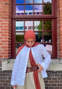 Witte button-down shirt over een beige jurk, aangevuld met een roestkleurige hijab en een bruine leren riem. Achtergrond van een bakstenen muur.