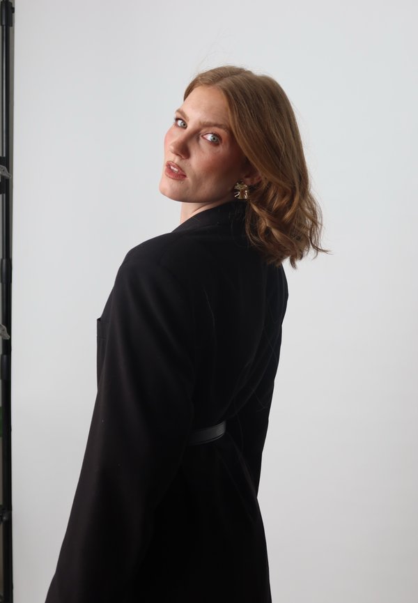 Black blazer with a smooth texture, featuring a belted waist and front pocket. Soft waves in hair frame the face against a white background.