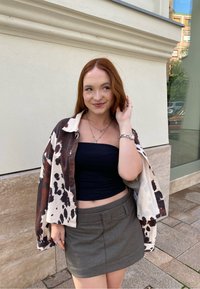 Kortærmet brun og beige ko-mønster skjorte over en sort crop top, parret med en grå mini-skort. Viser et afslappet, lagdelt look.