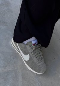 Sapatilha de camurça cinza com o logo da Nike em branco, parte superior texturizada e sola branca em contraste; emparelhada com calças escuras e meias claras.