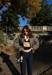 Hnědo-černá houndstooth bunda, béžový a námořnický pruhovaný svetr, tmavé džíny, držící černo-bílou vzorovanou tašku.