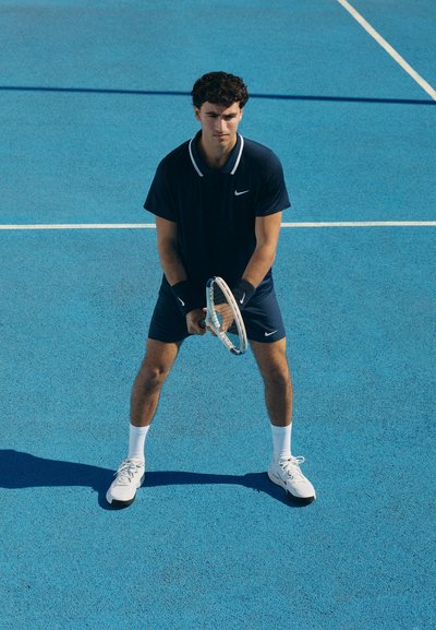 Camiseta deportiva azul marino con cuello blanco, pantalones cortos a juego, sosteniendo una raqueta de tenis. Zapatillas y calcetines blancos en una pista de tenis azul.