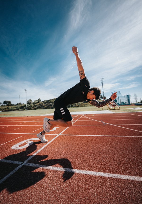 Athlète aux bras tatoués en tenue de sport noire sprintant depuis le starting-block sur une piste extérieure sous un ciel bleu clair.