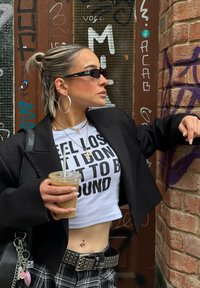 Sacou oversized negru, cămașă albă scurtă cu text îndrăzneț, fustă cu pattern carourile alb-negru, cercei cercuri și ochelari de soare. Ținând o cafea rece.