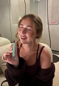 Rossetto verde menta in un tubetto a rotazione con dettagli argentati, tenuto da una persona che indossa un pullover scuro, sorridendo con gli occhi chiusi.