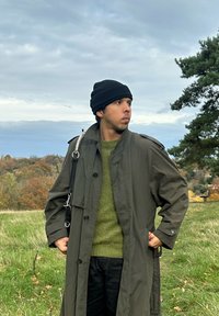 Man die een zwarte muts, olijfgroene trui en een lange donkergroene jas draagt, staat in een grasveld met bomen en een bewolkte lucht op de achtergrond.