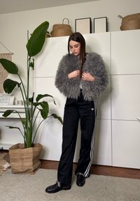 Femme ajustant une veste grise et duveteuse, portant un pantalon de survêtement noir avec des rayures blanches, debout dans une pièce avec une grande plante verte et des paniers tressés.