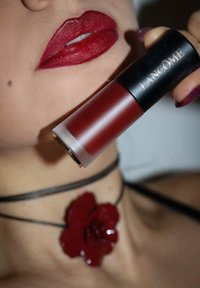 Lancôme huulipuna sylinterimäisessä pakkauksessa, jossa on musta kansi ja huurrettu runko; syvänpunainen väri huulilla; teksturoitu musta kukkaketju.