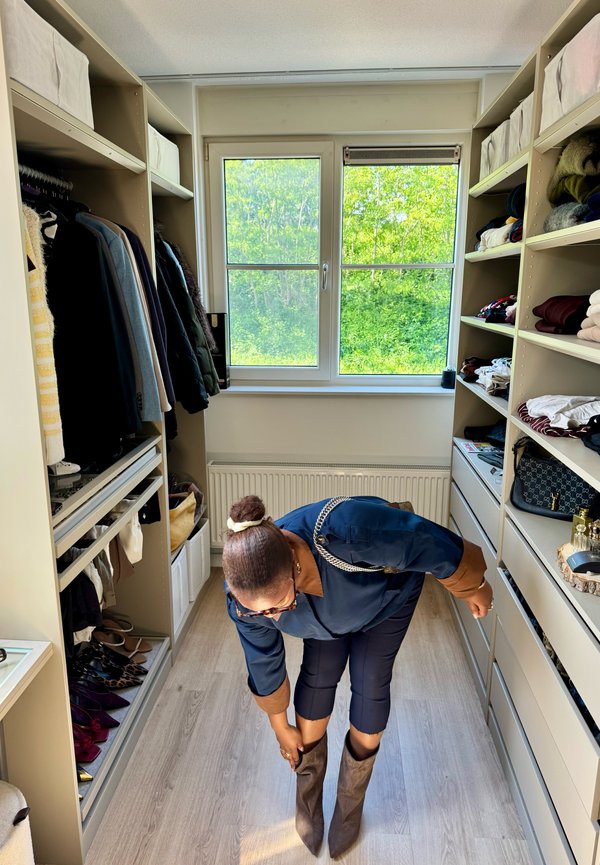 Femme portant une tenue bleu marine se penche pour ajuster des bottes marron montant jusqu'aux genoux dans un dressing avec des vêtements et des accessoires sur les étagères.