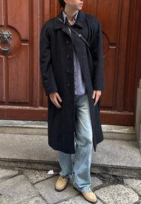 Homme portant un long manteau noir, une chemise à carreaux, un jean bleu clair, des chaussures beige et un sac bandoulière noir, debout sur des marches en pierre devant des portes en bois.