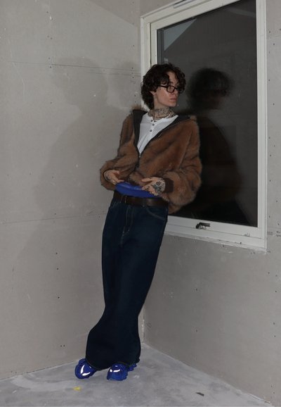 Chaqueta de piel sintética marrón con capucha, camisa blanca, cinturilla azul de Calvin Klein, vaqueros anchos de pierna oscura y zapatillas azules. Fondo gris liso.