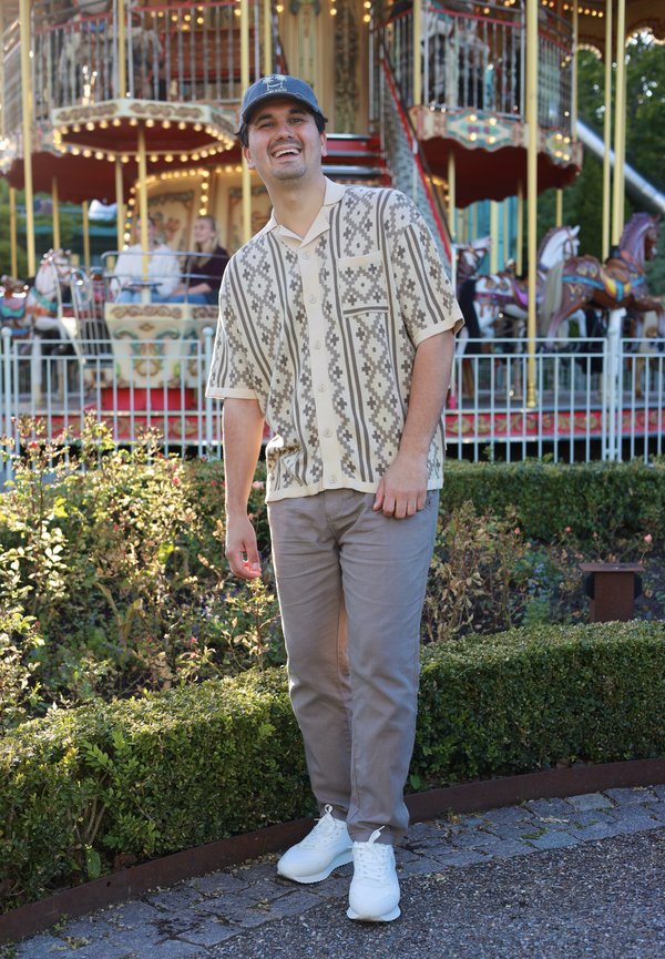 Mann lächelt und steht neben Büschen vor einem beleuchteten Karussell mit Menschen und Pferden in einem Freizeitpark oder auf einem Jahrmarkt.