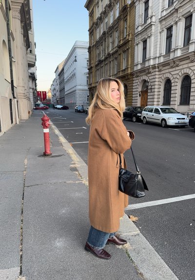 Abrigo de lana marrón, vaqueros azules y botines burdeos. Bolso negro de hombro. Escena urbana con edificios vintage y una boca de incendios.