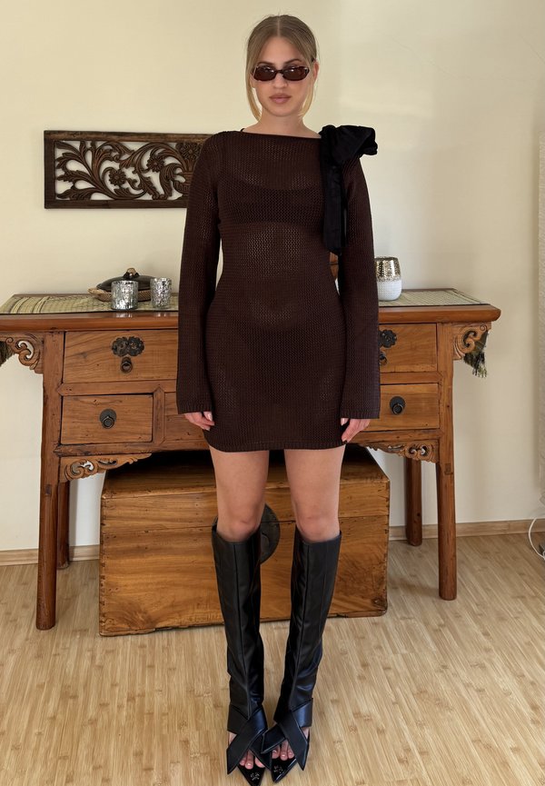 Vestido mini de punto marrón con mangas largas y lazo en el hombro, combinado con botas altas negras de puntera afilada y detalle de nudo.