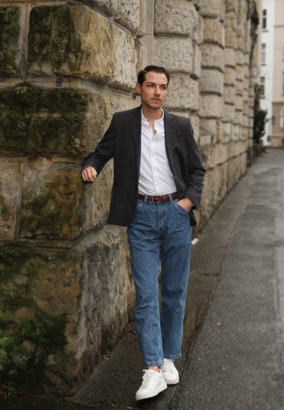 Hombre vistiendo un blazer gris oscuro sobre una camisa blanca, jeans azules y zapatillas blancas, de pie contra una pared de piedra en una calle mojada.