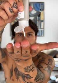 Man met tattoos die crème uit een tube op zijn open hand aanbrengt in een heldere kamer met een mirror en planken op de achtergrond.