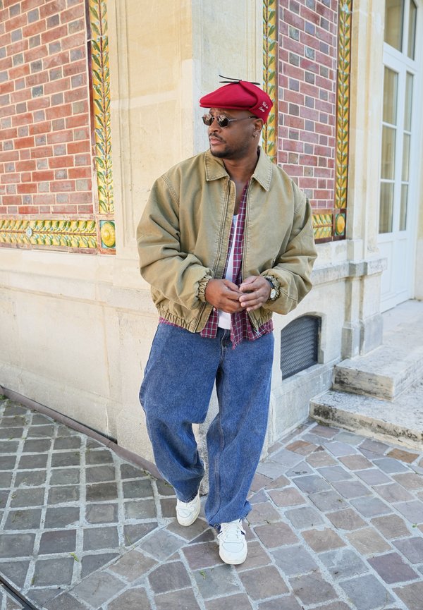 Uomo che indossa un berretto rosso, occhiali da sole, giacca beige, jeans blu larghi e scarpe da ginnastica bianche, in piedi vicino a un edificio con muri in mattone e pietra decorati.