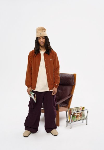 Chaqueta de pana marrón, gorro beige, camisa crema, pantalones cargo morados, zapatos de lona crema. De pie junto a un soporte para revistas y una silla.