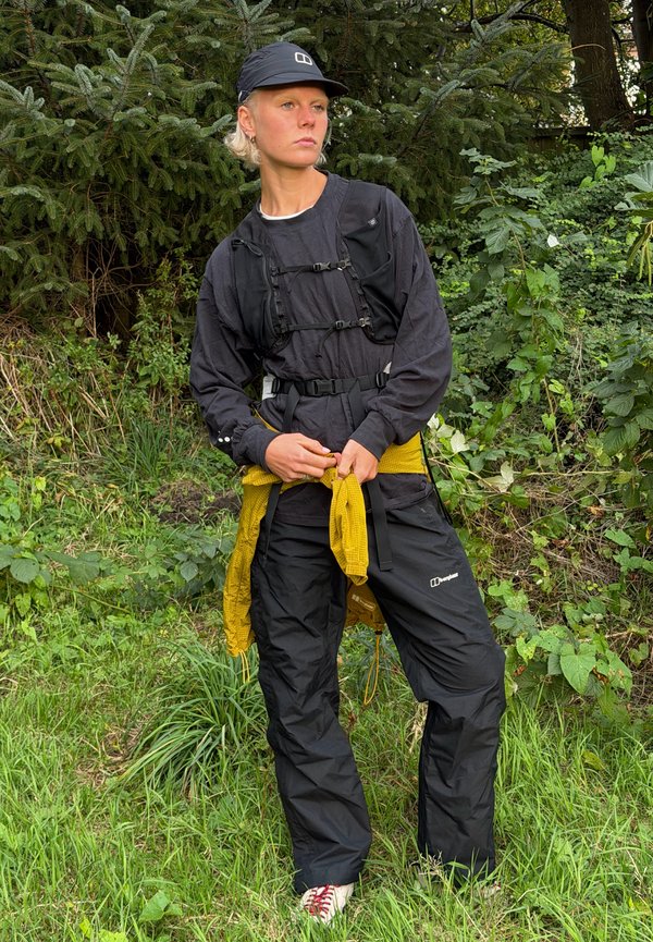 Una persona vestida con ropa negra de exterior y una gorra está entre una vegetación frondosa, sujetando una chaqueta amarilla alrededor de la cintura.