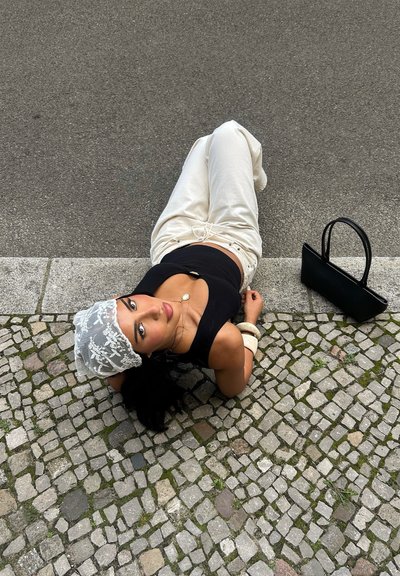 Mujer tumbada sobre un pavimento de adoquines, vestida con una camiseta sin mangas negra, pantalones deportivos blancos y un pañuelo de encaje en la cabeza, con un bolso negro cerca.