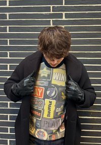 Fekete bőrkesztyűt és kabátot viselő személy, aki egy színes kötött pulóvert néz lefelé, amelyen szöveges és grafikás patchek találhatók, téglafal előtt.