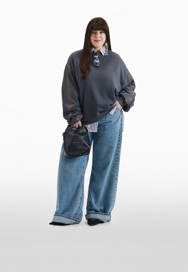 Kvinne med langt brunt hår iført stor, mørk grå genser, vide blå jeans, svarte sko, holder svart håndveske og solbriller på kragen.