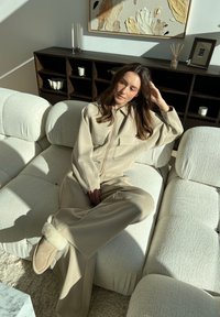 Beige loungewear set comprising a zip-up jacket with two front pockets and flared trousers. Made from soft fabric, paired with slippers on a textured sofa.