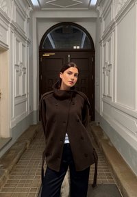 Brązowy dwurzędowy płaszcz z naramiennikami, z wysokim kołnierzem i miękką fakturą, zestawiony z ciemnymi spodniami w korytarzu.