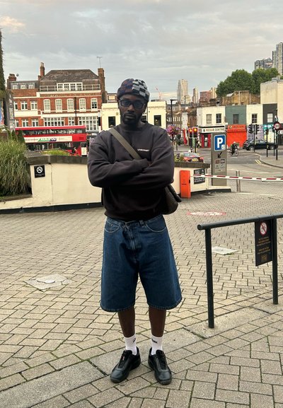 Hombre vestido con un sudadera oscura con texto, pantalones cortos de mezclilla anchos y zapatillas negras. Lleva un gorro con estampado y calcetines blancos. Fondo urbano.