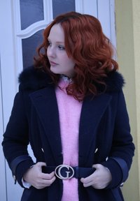 Jeune femme aux cheveux roux bouclés portant un manteau bleu marine à col en fourrure et un pull rose, attachant une ceinture noire avec une boucle dorée en forme de G.