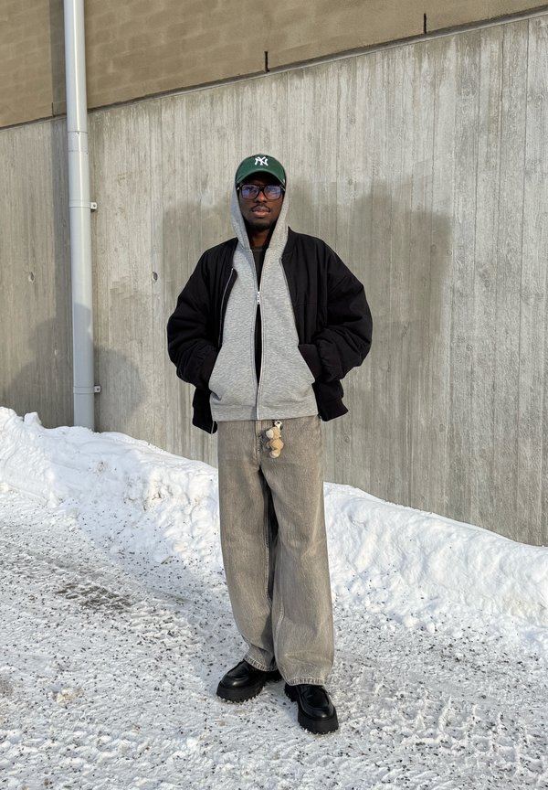 Hombre con chaqueta negra, sudadera con capucha gris, pantalones anchos grises, zapatos negros y gorra verde, de pie sobre terreno nevado junto a una pared de concreto.