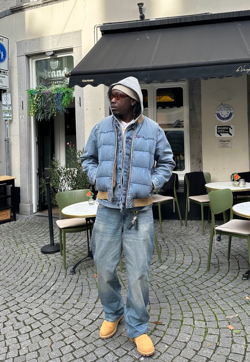 Τζάκετ puffer από τζιν επάνω από τζιν μπουφάν, γκρι φούτερ από κάτω, χαλαρά μπλε τζιν και καφέ μπότες. Ορατές λεπτομέρειες γυαλιών ηλίου και μπρελόκ.