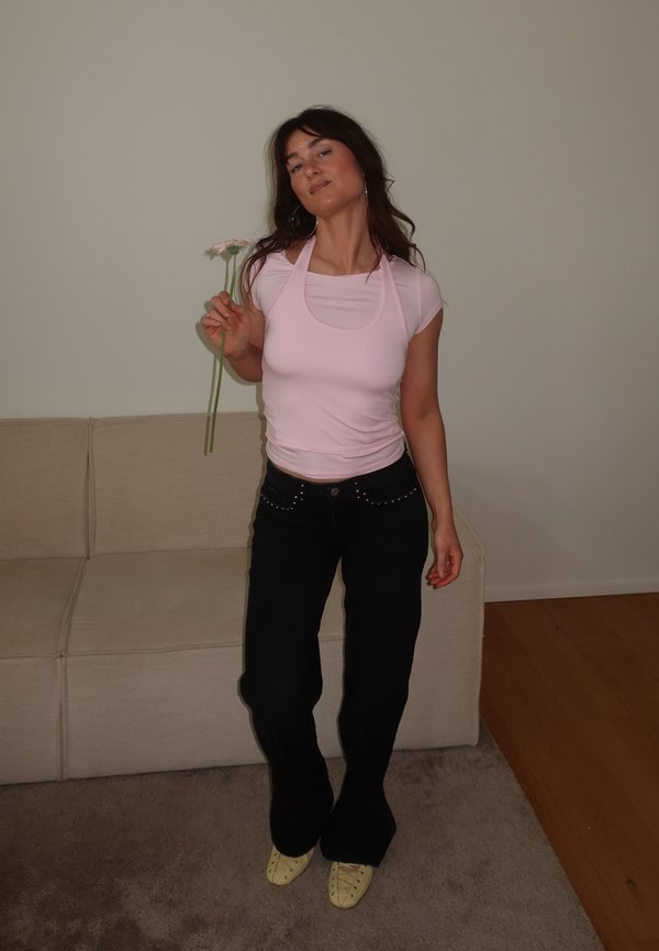 Mujer con una blusa rosa y pantalones negros, de pie junto a un sofá beige, sosteniendo dos flores rosas en su mano derecha en el interior.