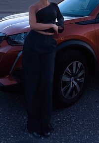 Top cropped preto de um ombro só combinado com calças largas pretas. O tecido parece suave. Ao fundo, um carro laranja é visível.