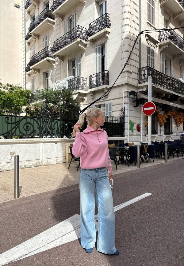 Maglione lavorato a maglia rosa chiaro con colletto alto, abbinato a jeans a gamba larga blu chiaro. In piedi su una strada vicino a un edificio con dettagli in ferro battuto.