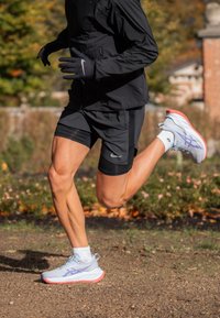 Világoskék és rózsaszín sportcipők; fekete rövidnadrágot és dzsekit visel. Futó félúton egy kavicsos úton, mögötte lombos háttér.