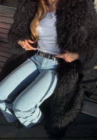 Personne portant un manteau noir duveteux, une chemise blanche, un jean bleu clair et une ceinture noire, assise sur un banc en bois avec les mains pointant vers la ceinture.