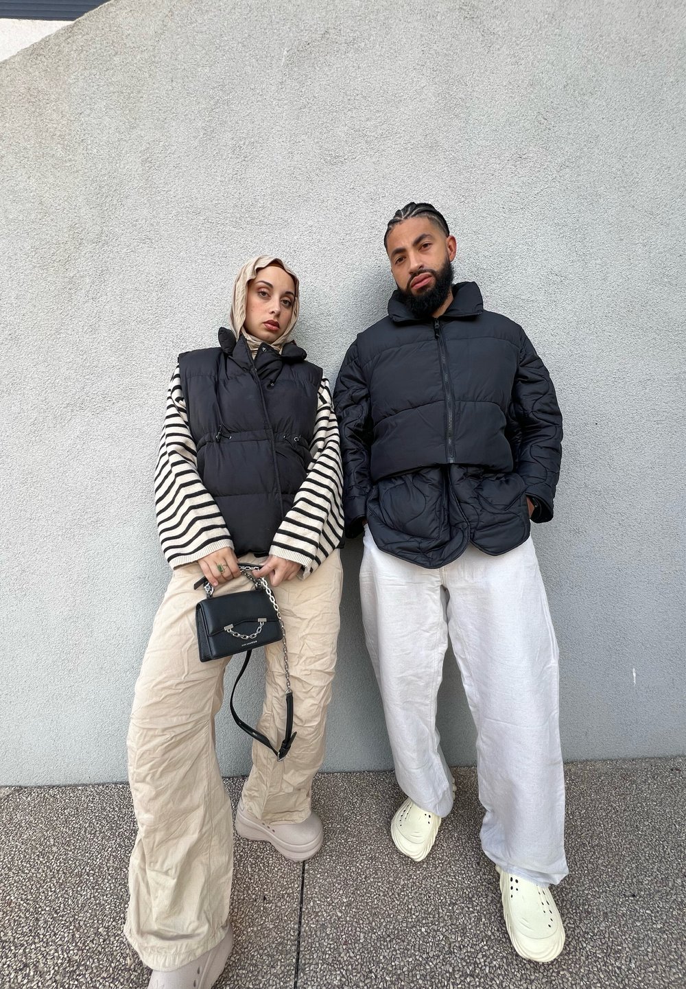 Δύο άτομα ποζάρουν σε έναν ανοιχτό γκρι φόντο. Φορούν μαύρες φουσκωτές ζακέτες. Η γυναίκα έχει μια ριγέ μπλούζα με μπεζ παντελόνια και πλατφόρμες.