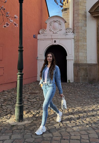 Mujer con chaqueta vaquera y vaqueros caminando por una calle empedrada junto a una puerta arqueada y ornamentada con el número 9, llevando un bolso blanco.
