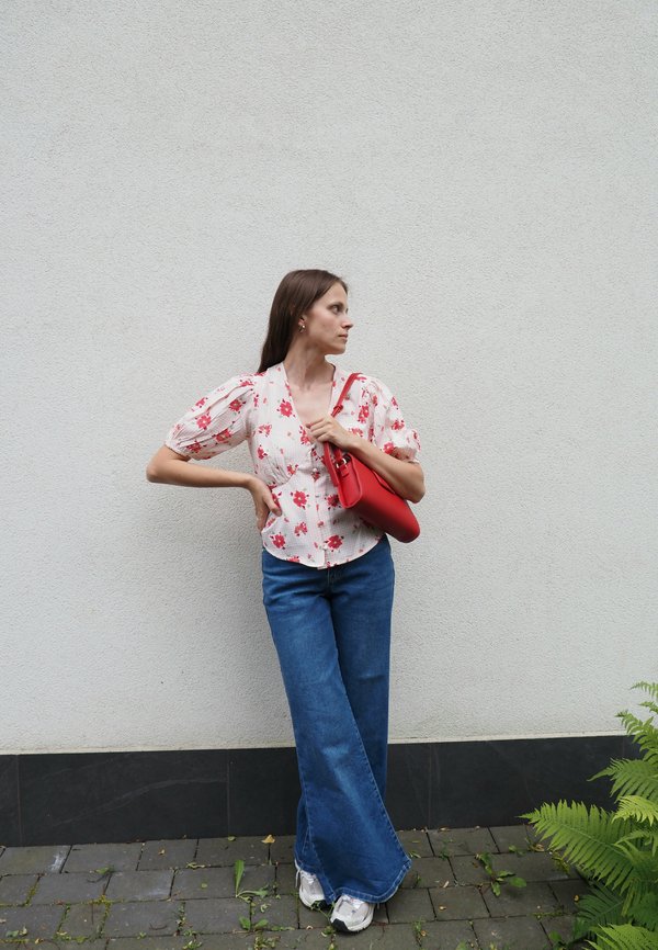 Blusa floreale bianca con motivi rossi, abbinata a jeans a gamba larga blu e sneakers bianche. Tiene in mano una borsa strutturata rossa.