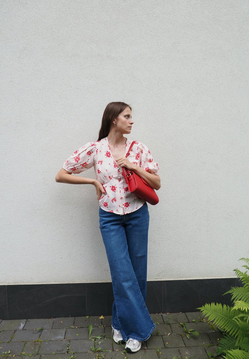 Weisse Bluse mit floralem Muster in Rot, kombiniert mit weit geschnittenen blauen Jeans und weißen Sneakern. Hält eine rot strukturierte Handtasche.