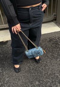 Borsa in denim con design trapuntato, colore azzurro chiaro, tracolla a catena dorata, tenuta da una persona che indossa jeans neri a vita alta e scarpe nere.