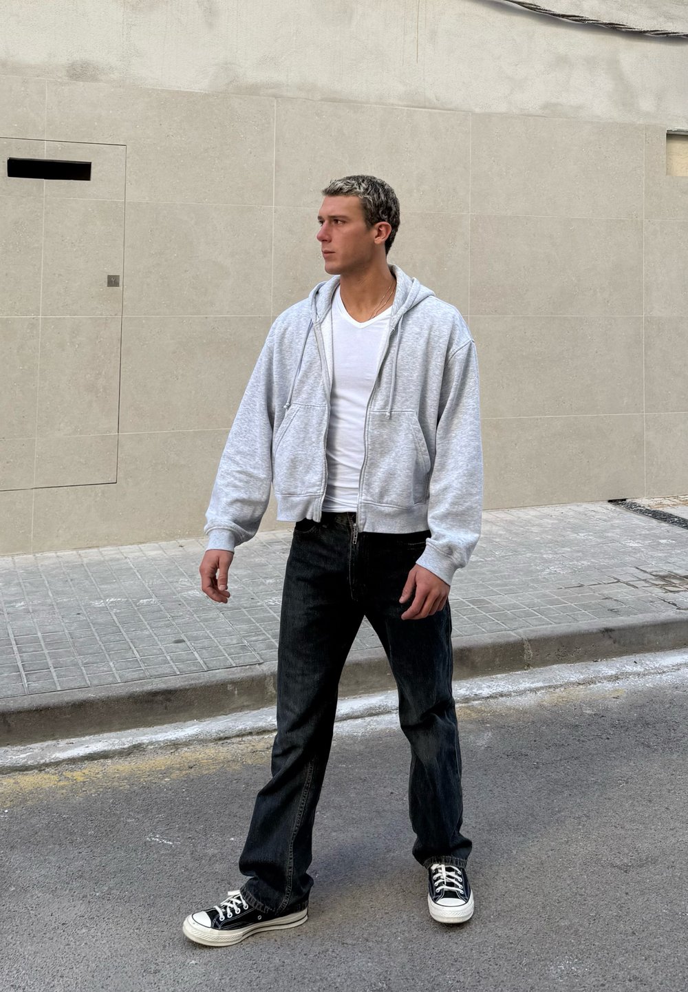 Moški v beli majici s kratkimi rokavi, sivem puloverju z zadrgo, črnih kavbojkah in črnih supergah stoji na ulici ob bež keramični steni in gleda levo.