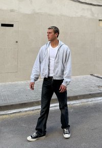 Moški v beli majici s kratkimi rokavi, sivem puloverju z zadrgo, črnih kavbojkah in črnih supergah stoji na ulici ob bež keramični steni in gleda levo.