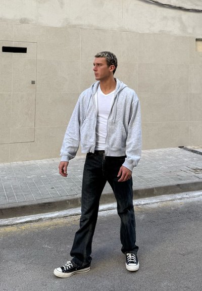 Hombre con camiseta blanca, sudadera con capucha gris con cremallera, vaqueros negros y zapatillas negras, de pie en una calle junto a una pared beige con azulejos, mirando hacia la izquierda.