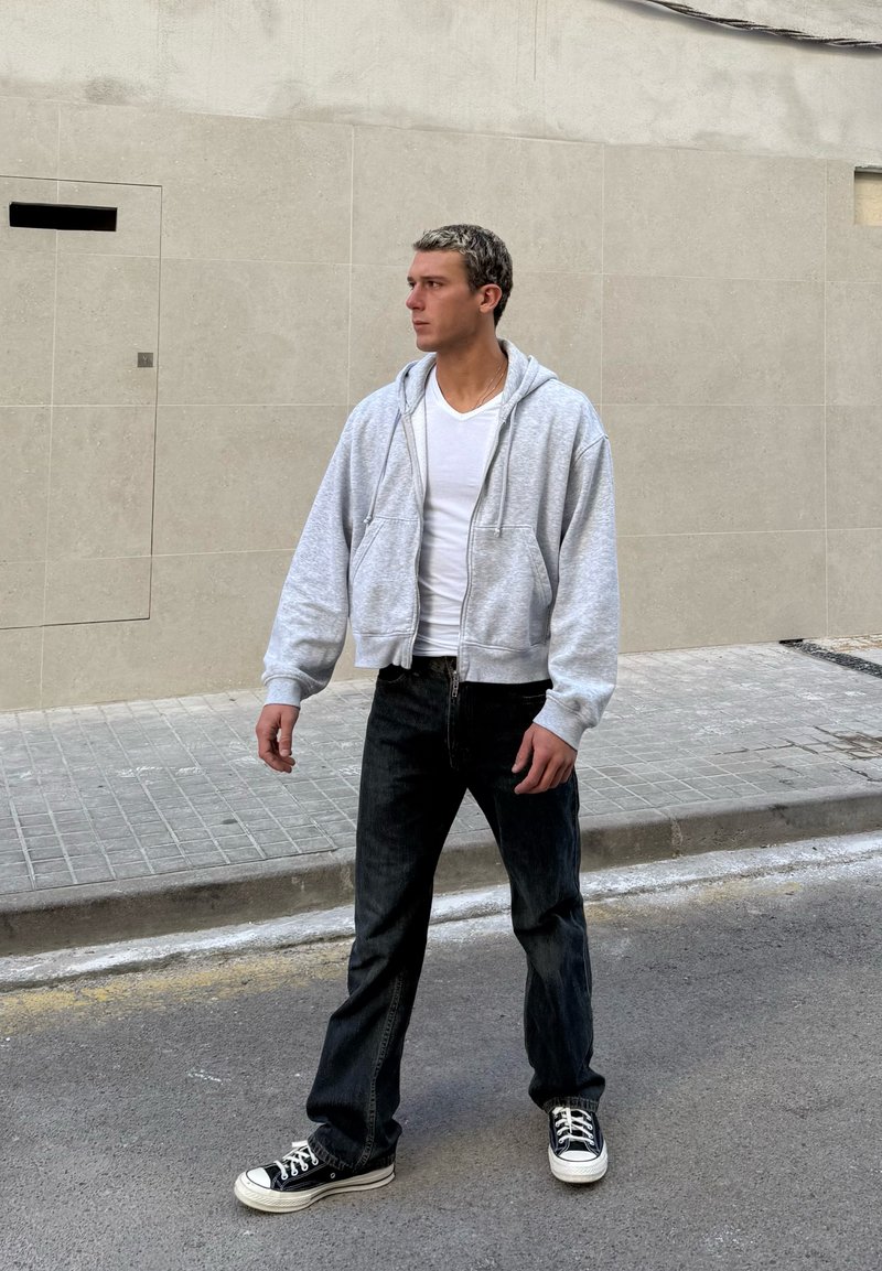 Man i vit t-shirt, grå zip-up hoodie, svarta jeans och svarta sneakers står på en gata vid en beige kakelvägg och tittar åt vänster.