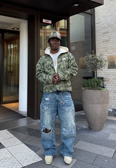 Chaqueta con estampado de camuflaje y capucha, combinada con jeans holgados y desgastados. Entre los accesorios notables se incluyen cadenas y una gorra con estampado.