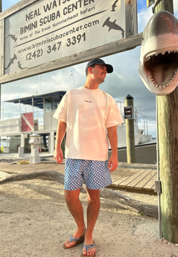 Férfi hétköznapi nyári ruhában, aki egy kikötő mólón áll, egy Neal Watson Bimini Scuba Center feliratú tábla alatt, amely mellett egy nagy cápa fej szobor található.