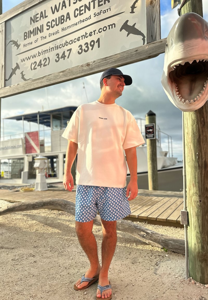 Vīrietis gadījuma vasaras apģērbā stāv pie jahtu piestātnes, zem zīmes "Neal Watson Bimini Scuba Center", kur redzama liela haizivju galvas skulptūra.