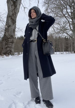 Mujer con abrigo de invierno, bufanda, pantalones anchos grises y botas negras, de pie sobre la nieve cerca de árboles deshojados, con un bolso negro colgado del hombro.